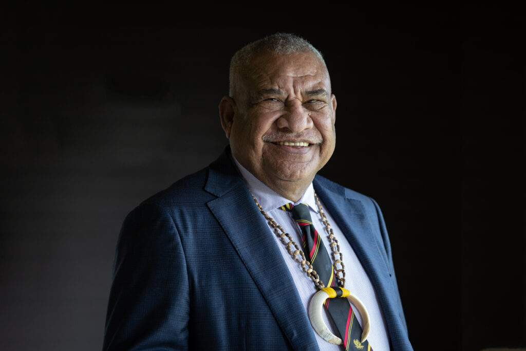 A photo of Femili PNG patron, John Kali. He is standing against a black background wearing a navy suit with a black, red and yellow tie. He is wearing a bore tusk necklace.
